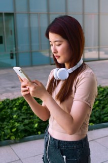 A young woman with headphones around her neck is focused on her smartphone. She stands outdoors