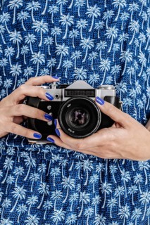 A woman in a blue dress with palm tree patterns is holding a vintage camera. Her fingernails are
