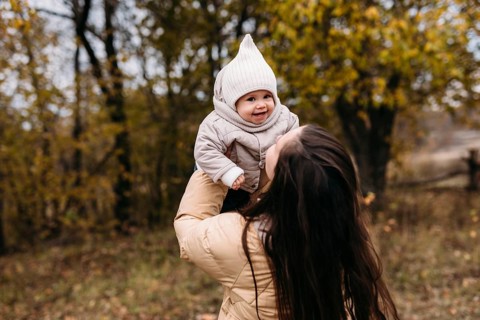 A mother joyfully lifts her warmly dressed child during a family walk in an autumn forest. The