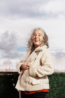 A joyful woman with white hair and a warm jacket stands on her balcony, smiling confidently at the