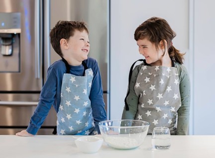 Two siblings wearing star patterned aprons smile at each other in a kitchen. A mixing bowl with