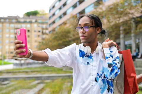 A confident man wearing headphones takes a selfie with a pink phone and shopping bags. He is