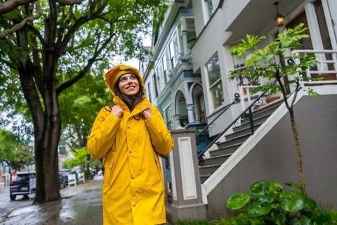 Young woman in a bright yellow raincoat with hood up, looking up and smiling while exploring a wet
