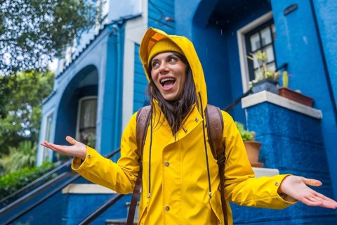 Woman in a yellow raincoat standing happily outside a vibrant blue house, smiling broadly and