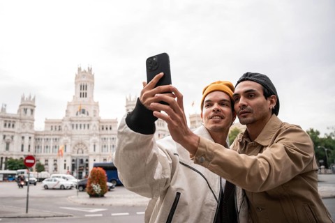 A gay couple takes a selfie in front of a historic Madrid building. They are dressed warmly and