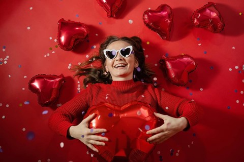 A joyful young woman lies on a red background surrounded by red heart-shaped balloons, holding a