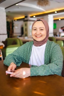 A smiling Muslim woman in a hijab sits comfortably at a table indoors, holding a smartphone. She
