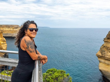 A woman with tattoos, wearing sunglasses, stands on a coastal cliff side overlooking the ocean in