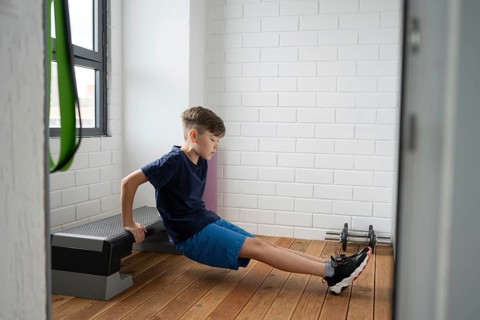 Young boy performs a seated dip exercise on a fitness bench indoors. The room has a modern design