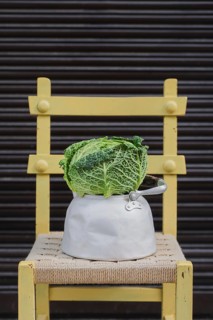 A fresh green cabbage sits in a rustic aluminum pot, placed on a woven seat of a yellow wooden