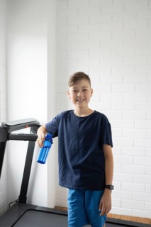 A young boy stands confidently by a treadmill, holding a blue water bottle. Dressed in casual