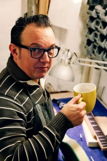 A skilled bass guitar luthier takes a coffee break in his cozy workshop. Surrounded by tools and