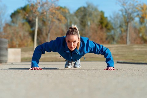 A woman in a blue jacket performs a push ups on a concrete surface outside, focused and determined.