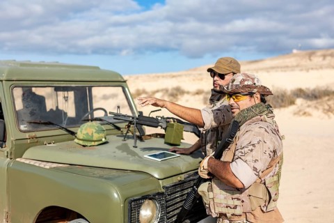 Two soldiers in camouflage uniforms discuss strategy next to a military vehicle in a desert setting