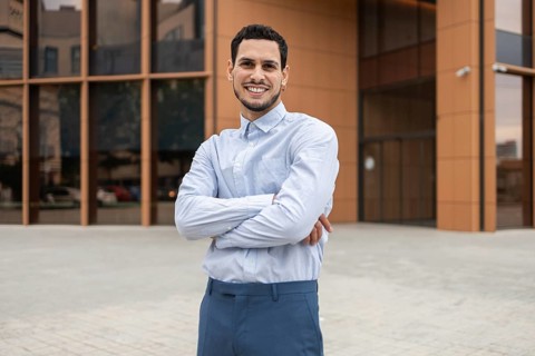 A young professional in business casual attire stands confidently with arms crossed, smiling in
