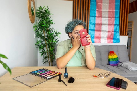 A transgender individual with blue hair is caught applying makeup in a mirror. Surrounded by a