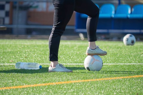 A person wearing sneakers and black pants stands with one foot on a soccer ball on a grass soccer