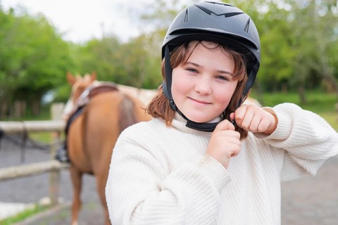 A young girl wearing a protective helmet fastens the strap, ready to enjoy a horse ride on a sunny