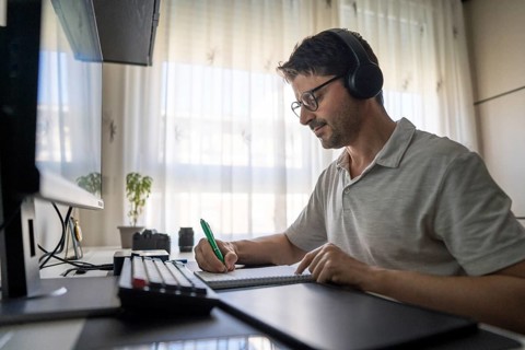 A man sits at his desk wearing headphones, focused on learning Mandarin online. He takes notes in a