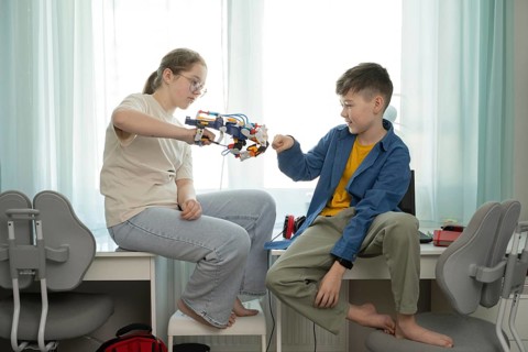 Two siblings engage with a mechanical toy arm in a bright bedroom. The girl operates the toy, while