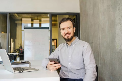 A man in a light shirt sits at a desk with a smartphone. A laptop and notepad accompany him in a