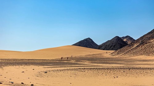 Vast desert landscape with dunes, mountains and small camel caravans under a blue sky, The endless
