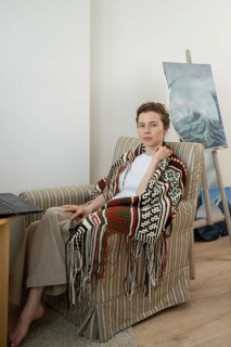 A woman wearing a patterned shawl sits in a cozy armchair beside a wooden table with a laptop. A