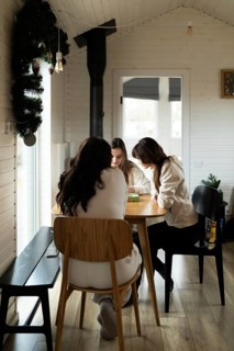 Three young women engage in a cozy indoor gathering, seated at a wooden table in a warmly decorated
