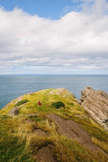 A female hiker in a vibrant red dress ascends a lush, rocky hillside at Signal Hill, Newfoundland &