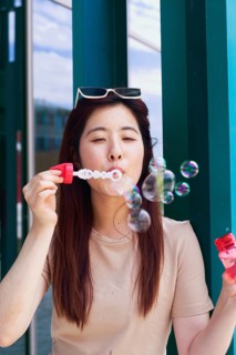 A young woman joyfully blows bubbles outside, capturing a playful moment. She wears sunglasses and