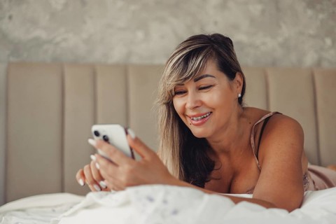 A smiling woman enjoys browsing her smartphone while lying comfortably on a bed. Her relaxed