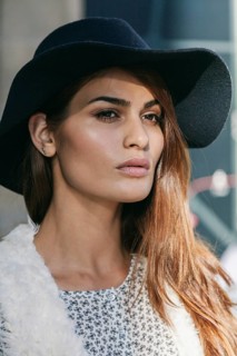 A woman wearing a wide-brimmed hat and a patterned blouse gazes thoughtfully. Her long hair falls