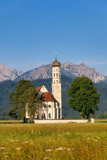 Church in the middle of a green meadow, surrounded by trees, with towering mountains in the