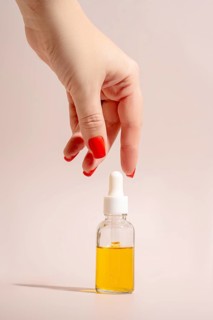 A hand with red manicured nails reaches for a clear glass dropper bottle filled with golden