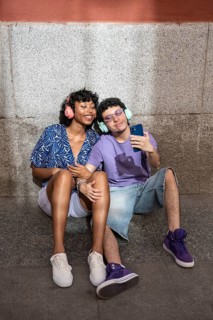 A joyful multiethnic LGBTQ+ couple sitting together, wearing headphones and sharing a smartphone,