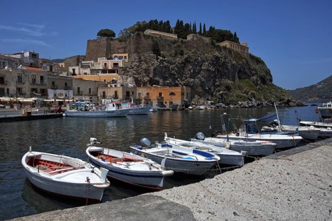 Boats are lying in the water of a harbor in front of a rocky mountain face and old buildings,
