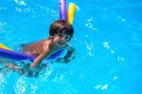 Adorable young child holding colorful pool noodles, swimming and smiling in bright blue water on a
