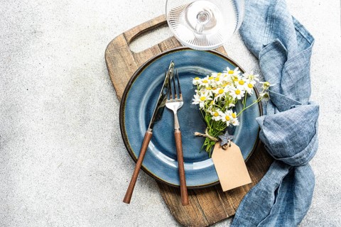 Top view of a summer daisy bouquet on a blue ceramic plate with a wooden cutting board, silverware,