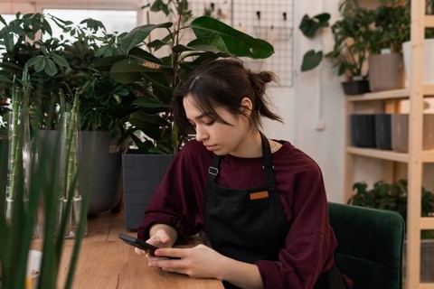 Young woman in a black apron sits at a wooden table, using her phone. Surrounded by lush green