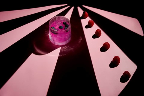 A vibrant pink cocktail in a glass, garnished with blueberries, is set against a striking pink-lit