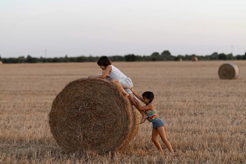 Two young siblings enjoy climbing and playing on a large hay bale in a field at dusk, showcasing