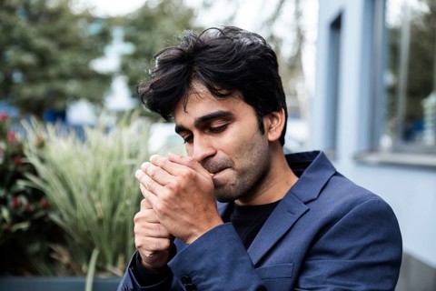 A indian man wearing a blue jacket is lighting a cigarette while standing outdoors. The background