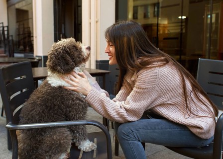 A young woman with long hair enjoys a moment with her dog at an outdoor cafe. She lovingly holds