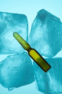 A glass ampoule containing yellow liquid rests on blue ice cubes against a light background,