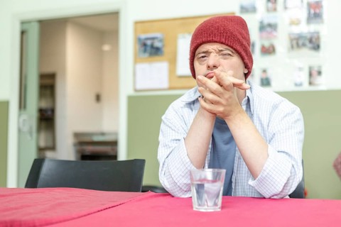 A man in a mental health residence sits contemplatively at a table, wearing a red beanie and casual