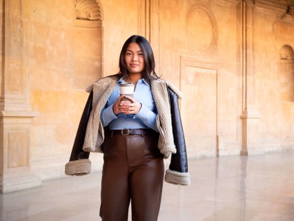 A young woman holding a coffee cup stands at an ornate historical building. She wears fashionable