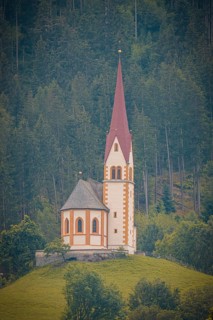 Church with pointed red tower on a hill surrounded by dense forest and mountains, Mayrhofen,