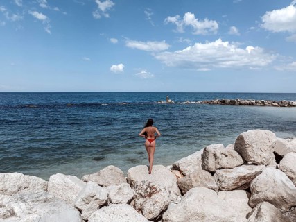 A young woman stands on the rocky shores of an Italian seaside, admiring the vast expanse of the