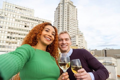 Two cheerful friends pose for a selfie while holding glasses of white wine on a rooftop The city