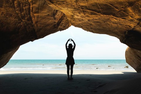A young Latin woman stands under a rocky arch in Mancora, Peru, forming a heart shape with her